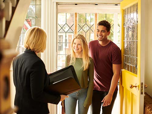 A young couple visiting a house that is for sale. An agent greent them, she holds a folder.